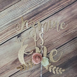 May include: Gold glitter cake topper with the text "Jasmine is one" and pink and green paper flowers.