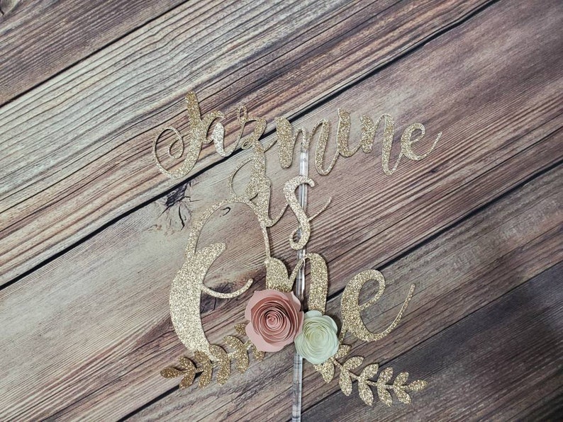 May include: Gold glitter cake topper with the text "Jasmine is one" and pink and green paper flowers.