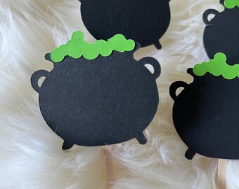 May include: Four black cauldron cupcake toppers with green frosting on top. The toppers are on a white fluffy surface.
