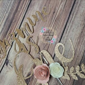May include: Gold glitter cake topper with the name "Joanne" and a floral design. The flowers are pink, white, and green.