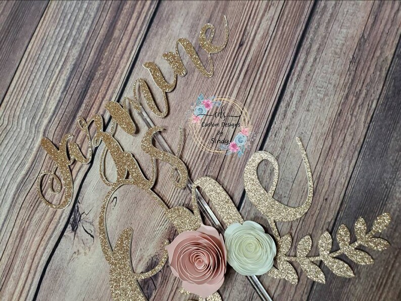 May include: Gold glitter cake topper with the name "Joanne" and a floral design. The flowers are pink, white, and green.