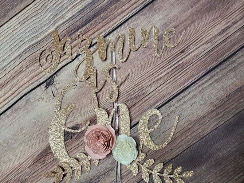 May include: Gold glitter cake topper with the words "One is the" and a floral design. The flowers are pink and white.