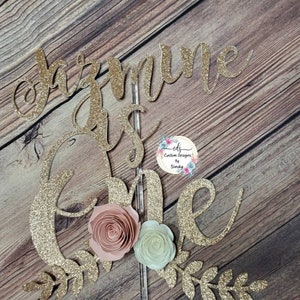May include: Gold glitter cake topper with the words "Jazmine is one" and a floral design. The topper is perfect for a first birthday party.
