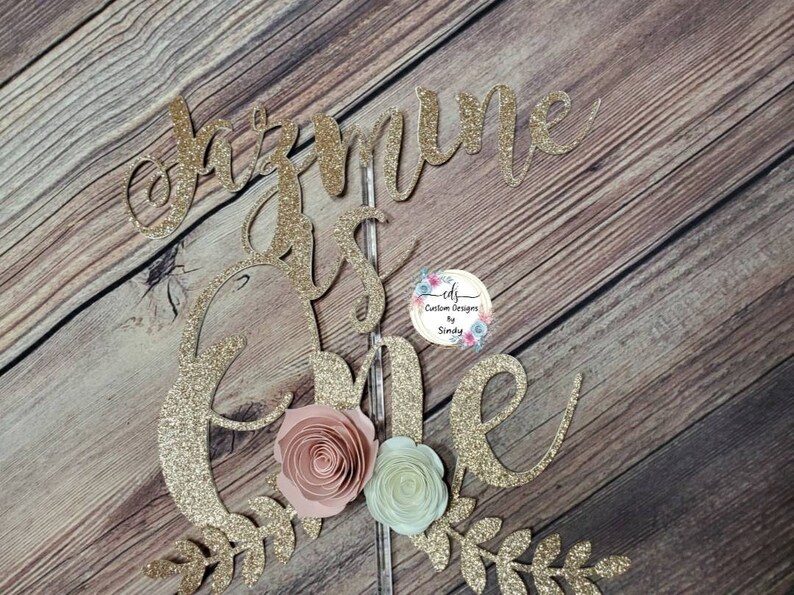 May include: Gold glitter cake topper with the words "Jazmine is one" and a floral design. The topper is perfect for a first birthday party.
