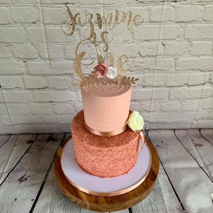 May include: A two-tiered cake decorated with pink frosting and gold sprinkles. The top tier has a simple pink frosting design, while the bottom tier is covered in gold sprinkles. The cake is topped with a gold glitter topper that says "Jazmine is One" and has small pink and white flowers.