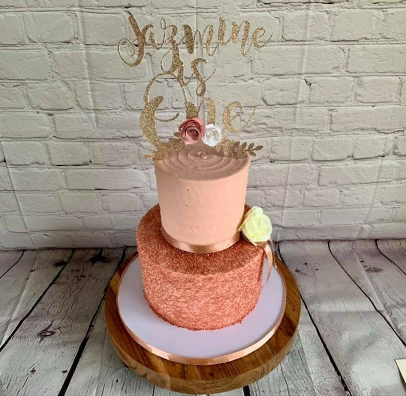 May include: A two-tiered cake decorated with pink frosting and gold sprinkles. The top tier has a simple pink frosting design, while the bottom tier is covered in gold sprinkles. The cake is topped with a gold glitter topper that says "Jazmine is One" and has small pink and white flowers.