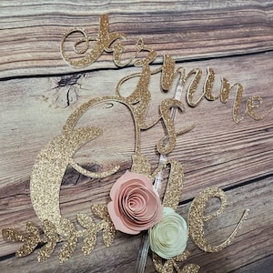 May include: Gold glitter cake topper with the words "One is the loneliest number" and a pink and white paper flower.