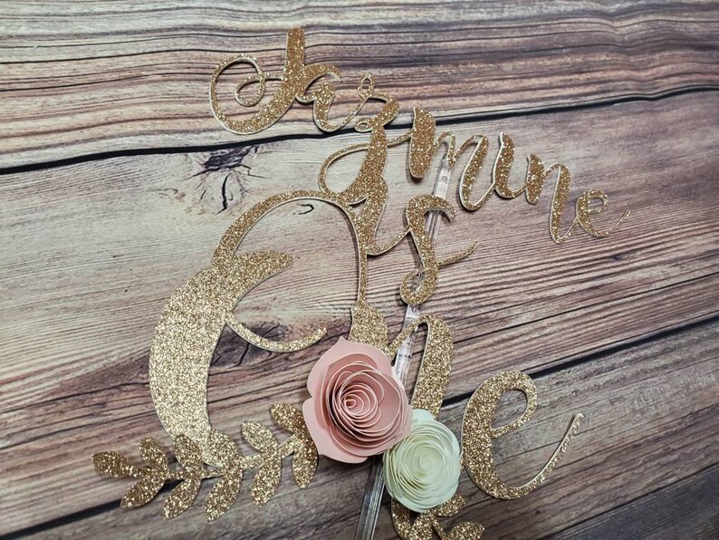 May include: Gold glitter cake topper with the words "One is the loneliest number" and a pink and white paper flower.
