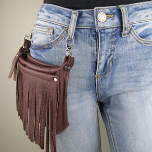 May include: A brown leather fringe bag with a silver chain and clip. The bag is worn on a person's waist.