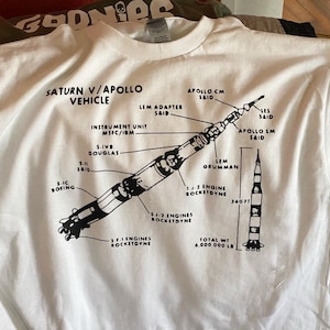 Puede incluir: Camiseta blanca con una ilustración en blanco y negro del cohete Saturno V, etiquetada con los nombres de las diferentes partes y fabricantes. El texto "Saturn V/Apollo Vehicle" está en la parte superior de la ilustración. El texto "Total Wt 6,000,000 LB" está en la parte inferior de la ilustración.