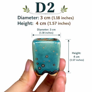 May include: A small square ceramic planter labeled D2 with a teal glaze and dark circular crystalline spots. It measures 1.18 inches in diameter and 1.57 inches in height. The base features a dark, uneven rim and a cutout design on the bottom edge.
