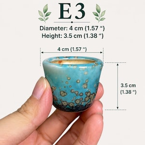 May include: A small ceramic pot with a turquoise and dark blue crystalline glaze featuring circular patterns. The pot is labeled E3 and measures 1.57 inches in diameter and 1.38 inches in height, as indicated by the text and dimension lines.