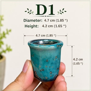 May include: A small, teal ceramic cup with a speckled glaze and a flared rim. The cup features the text D1 and dimensions of 1.85 inches in diameter and 1.65 inches in height, as indicated by the technical measurement lines shown in the image.