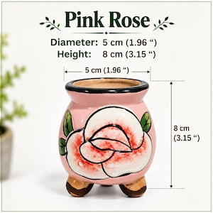 May include: A pink ceramic planter labeled Pink Rose with a hand-painted rose design. It features a black rim, three brown feet, and measures 1.96 inches in diameter and 3.15 inches in height as indicated by the technical text.
