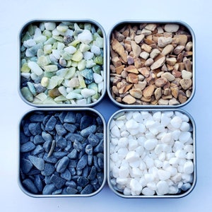 May include: Four small square containers filled with different types of decorative stones. The containers are made of metal and have a silver finish. The stones are white, black, green, and brown.