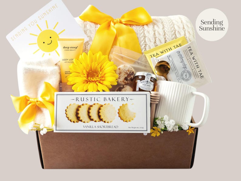 Peut inclure: Un panier cadeau garni de divers articles, dont une tasse blanche, des biscuits sabl&eacute;s &agrave; la vanille, des sachets de th&eacute;, une lotion pour le corps et une fleur jaune. Le panier est nou&eacute; avec un ruban jaune et contient une carte "Sending You Sunshine".