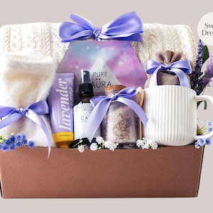Peut inclure: Un coffret cadeau de bien-&ecirc;tre avec une couverture blanche en tricot torsad&eacute;, une serviette blanche roul&eacute;e, de la lotion &agrave; la lavande, une huile essentielle, des sels de bain et une tasse blanche. Les articles sont li&eacute;s avec des rubans violets dans une bo&icirc;te marron. Le texte "Sweet Dreams" est visible.