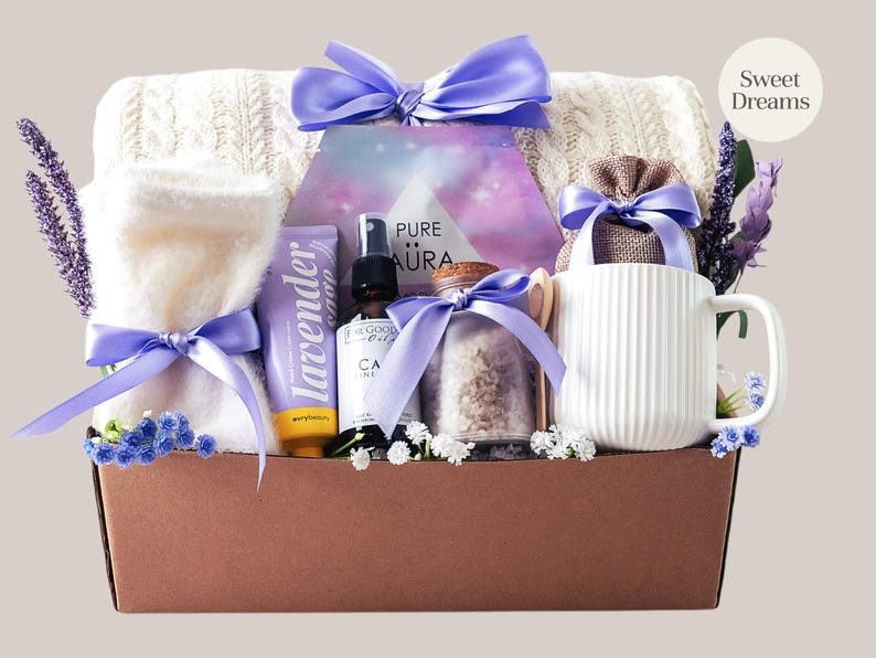 Peut inclure: Un coffret cadeau de bien-&ecirc;tre avec une couverture blanche en tricot torsad&eacute;, une serviette blanche roul&eacute;e, de la lotion &agrave; la lavande, une huile essentielle, des sels de bain et une tasse blanche. Les articles sont li&eacute;s avec des rubans violets dans une bo&icirc;te marron. Le texte "Sweet Dreams" est visible.