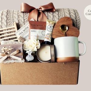 Peut inclure: Un panier cadeau garni d'articles r&eacute;confortants. Il comprend une couverture en tricot torsad&eacute; cr&egrave;me, une tasse blanche et marron, un dessous de verre en forme de c&oelig;ur en bois avec une cuill&egrave;re dor&eacute;e, du th&eacute;, du miel, une bougie, du savon et un n&oelig;ud d&eacute;coratif. Le texte "Cozy Comfort" est visible.