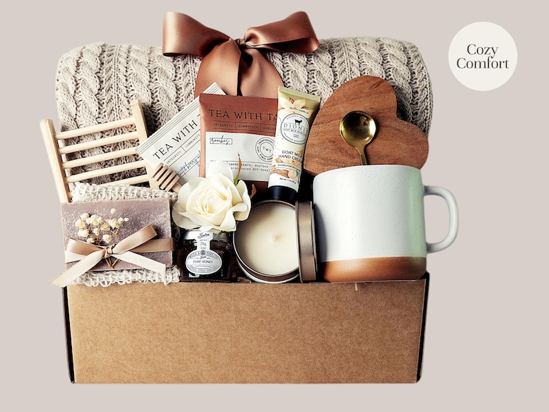 Peut inclure: Un panier cadeau garni d'articles r&eacute;confortants. Il comprend une couverture en tricot torsad&eacute; cr&egrave;me, une tasse blanche et marron, un dessous de verre en forme de c&oelig;ur en bois avec une cuill&egrave;re dor&eacute;e, du th&eacute;, du miel, une bougie, du savon et un n&oelig;ud d&eacute;coratif. Le texte "Cozy Comfort" est visible.