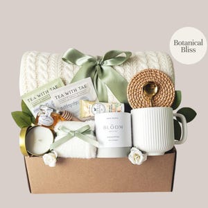 Peut inclure: Un panier cadeau rempli d'articles de bien-&ecirc;tre. Il comprend une couverture en tricot torsad&eacute; cr&egrave;me, des sachets de th&eacute;, du miel, une bougie, une tasse et un dessous de verre tress&eacute; avec une cuill&egrave;re dor&eacute;e. Le panier est nou&eacute; avec un ruban vert et contient le texte "Botanical Bliss".