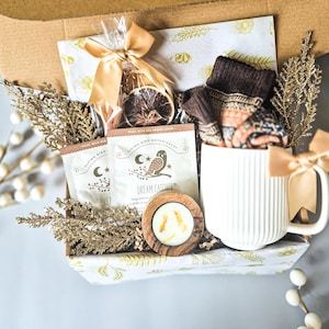 May include: A gift box filled with a white mug, socks, tea packets, a candle, and dried orange slices. The mug has vertical lines and a gold bow. The tea packets read "Dream Catcher" and feature an owl illustration.