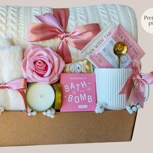 Peut inclure: Un panier cadeau avec une couverture en tricot torsad&eacute; cr&egrave;me, une tasse blanche, une rose rose et une bougie. Le panier comprend une bombe de bain rose, des sachets de th&eacute; et une cuill&egrave;re dor&eacute;e. Les articles sont nou&eacute;s avec des rubans roses. Le texte "Pretty in pink" est visible.