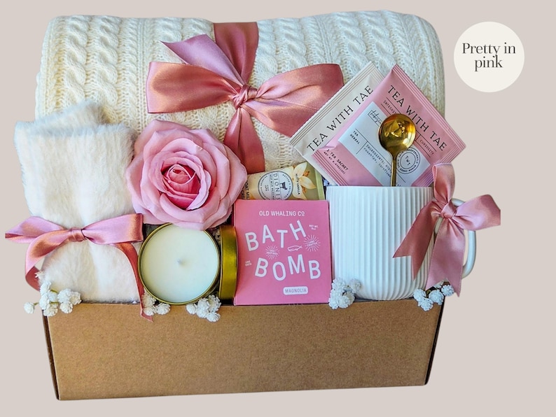 Peut inclure: Un panier cadeau avec une couverture en tricot torsad&eacute; cr&egrave;me, une tasse blanche, une rose rose et une bougie. Le panier comprend une bombe de bain rose, des sachets de th&eacute; et une cuill&egrave;re dor&eacute;e. Les articles sont nou&eacute;s avec des rubans roses. Le texte "Pretty in pink" est visible.
