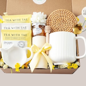 Peut inclure: Un coffret cadeau rempli de th&eacute;, de miel, d'une tasse et d'un dessous de verre tiss&eacute;. La bo&icirc;te contient des sachets de th&eacute; avec l'inscription "TEA WITH TAE", un petit pot de miel, une tasse blanche, un b&acirc;tonnet &agrave; miel et une bougie d&eacute;corative.