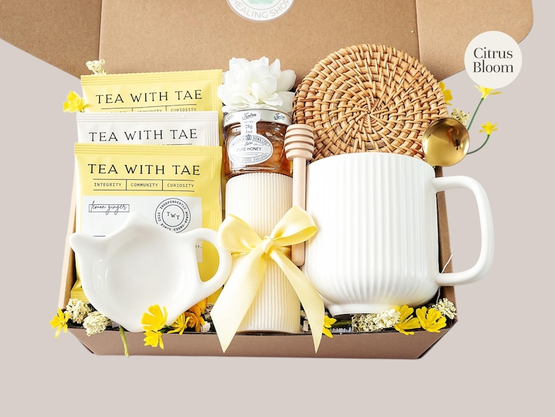 Peut inclure: Un coffret cadeau rempli de th&eacute;, de miel, d'une tasse et d'un dessous de verre tiss&eacute;. La bo&icirc;te contient des sachets de th&eacute; avec l'inscription "TEA WITH TAE", un petit pot de miel, une tasse blanche, un b&acirc;tonnet &agrave; miel et une bougie d&eacute;corative.
