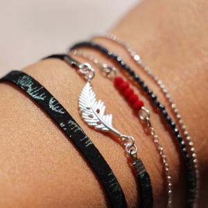Peut inclure: Un ensemble de bracelets superposés composé d'un bracelet à perles noires et argentées, d'un bracelet à chaîne argentée avec un pendentif en forme de plume et d'un bracelet en tissu noir à motif vert.