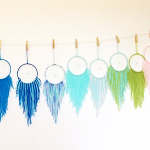 May include: A row of 14 dreamcatchers hanging from a string, each with a different color of yarn. The dreamcatchers are all white with a white web and a dark blue ring. The yarn colors are black, blue, teal, light blue, turquoise, green, light green, pink, red, orange, purple, and yellow.