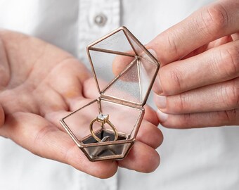 May include: A clear glass ring box with a gold metal frame, open and displaying a gold ring with a single diamond. The box is held in the palm of a hand.