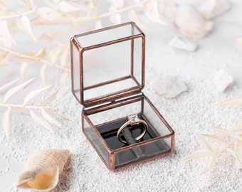 May include: A gold ring with a square-shaped diamond sits inside a clear glass ring box with a copper frame. The box is on a white surface with dried white branches and seashells.