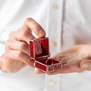 May include: A red velvet ring box with a gold trim, open to reveal a silver ring with a single diamond.