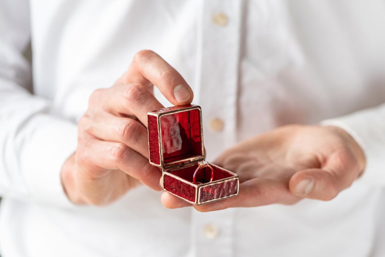 May include: A red velvet ring box with a gold trim, open to reveal a silver ring with a single diamond.