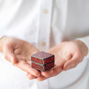 May include: A small, red, square-shaped ring box with a textured lid. The box is held in the open palms of a person.