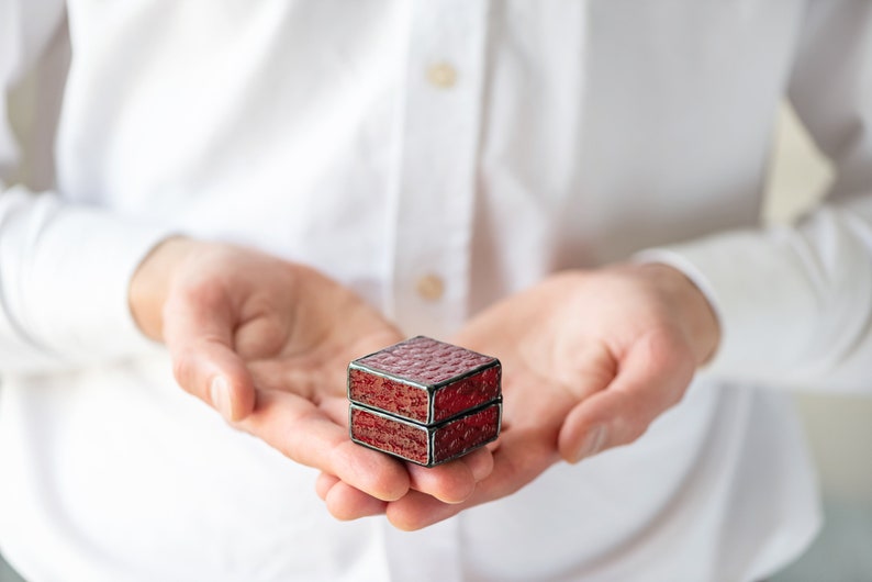 May include: A small, red, square-shaped ring box with a textured lid. The box is held in the open palms of a person.