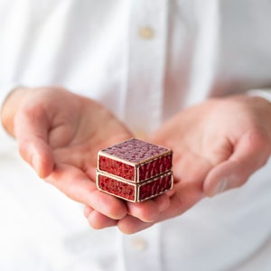 May include: A small, red, square-shaped box with a gold trim. The box is stacked on top of another similar box. The box is being held in the palm of a person's hand.