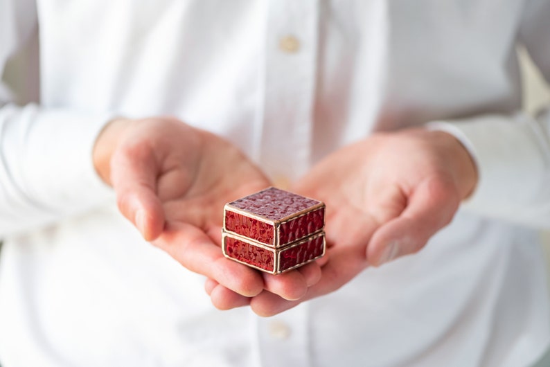 May include: A small, red, square-shaped box with a gold trim. The box is stacked on top of another similar box. The box is being held in the palm of a person's hand.