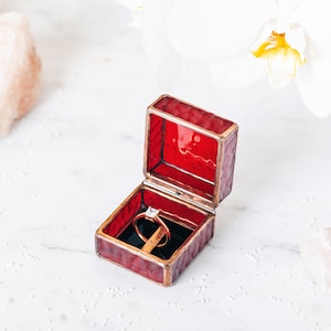 May include: A red velvet ring box with a gold trim is open, revealing a diamond engagement ring. The box is sitting on a white marble surface.