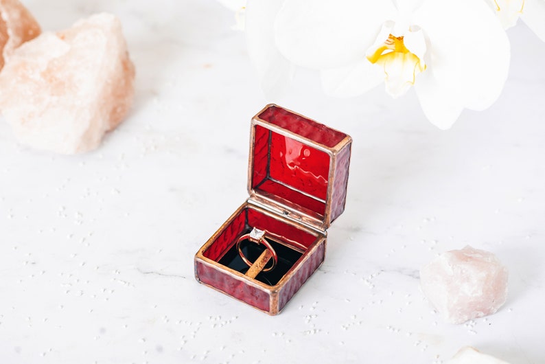 May include: A red velvet ring box with a gold trim is open, revealing a diamond engagement ring. The box is sitting on a white marble surface.