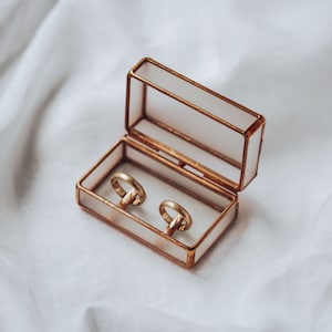May include: Two gold wedding bands in a clear glass ring box with a gold trim. The box is open and the rings are visible.