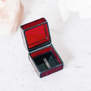 May include: A small, red, square-shaped ring box with a black velvet interior. The box is made of stained glass and has a hinged lid.