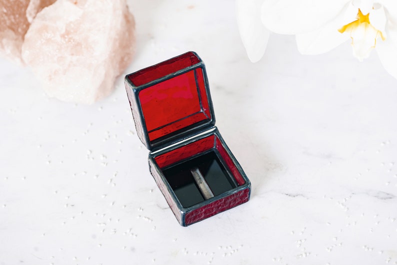 May include: A small, red, square-shaped ring box with a black velvet interior. The box is made of stained glass and has a hinged lid.