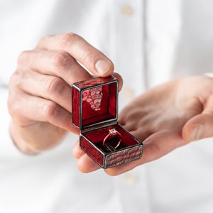 May include: A red velvet ring box with a gold band ring with a clear stone inside.