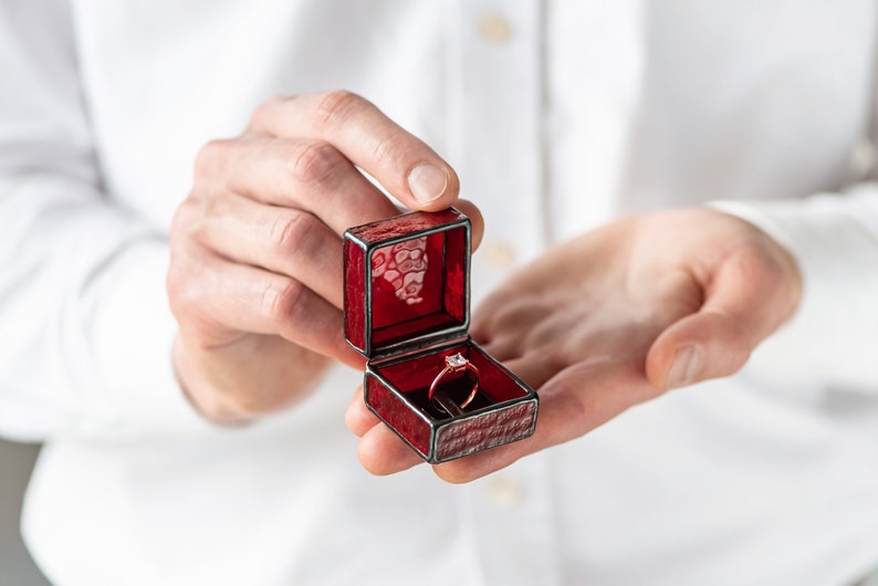May include: A red velvet ring box with a gold band ring with a clear stone inside.
