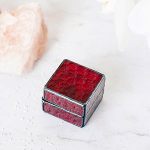 May include: A small, square, red glass ring box with a black border. The box is made of stained glass and has a textured surface.