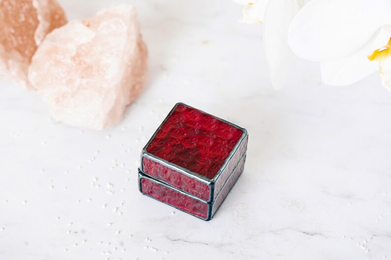 May include: A small, square, red glass ring box with a black border. The box is made of stained glass and has a textured surface.