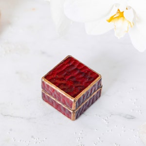 May include: A small, square, red ring box with a gold trim. The box is closed and sitting on a white surface.